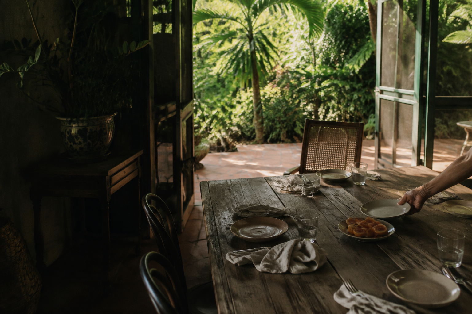 A hand sets a plate beside an empty cane chair on a veranda opening onto a green garden.