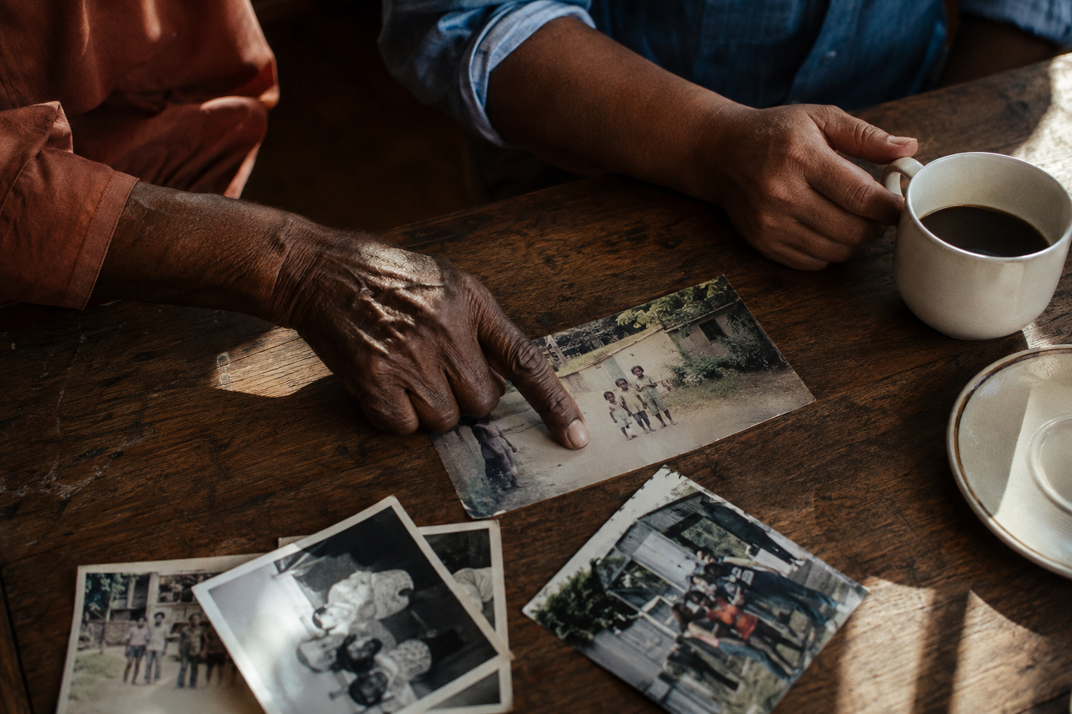 Two people sharing old family photographs at a wooden table, their hands beside a cup of coffee.